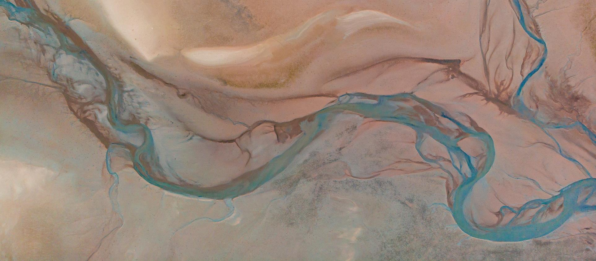 Aerial satellite view of a river meandering through desert dunes — teal channels threading through earth tones, evoking systems seen whole